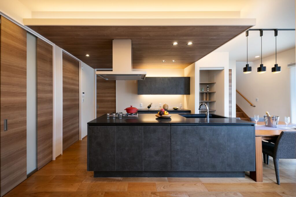 川口市の和モダンな注文住宅。木目天井の下がり壁×間接照明の組合せで上質感が漂い、空間に映えるダークカラーがおしゃれなオリジナルキッチン。